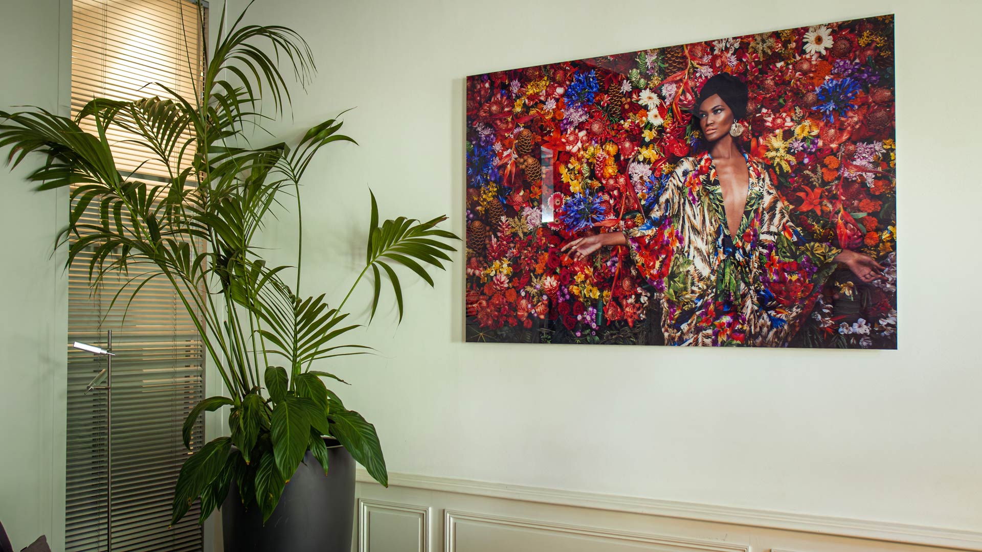 Art floral et végétation intérieure Tableau d’une femme entourée de fleurs colorées accroché au mur, à côté d’une grande plante verte en pot.